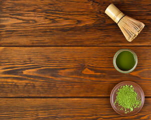 Green matcha tea in a cup with powdered matcha tea in a plate and bamboo matcha tea whisk also know as chasen on a wooden background. Top view. Text space.