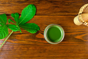 Matcha tea in a cup and green leaves with bamboo matcha tea whisk also know as chasen on a wooden background.