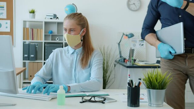 Woman in face mask and gloves is working with computer then waving good-bye to colleague in office. Pandemic healthcare and workplace concept.