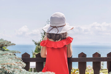 Coniferous coast. A little girl in a white beach hat and red dress stands with her back to the...