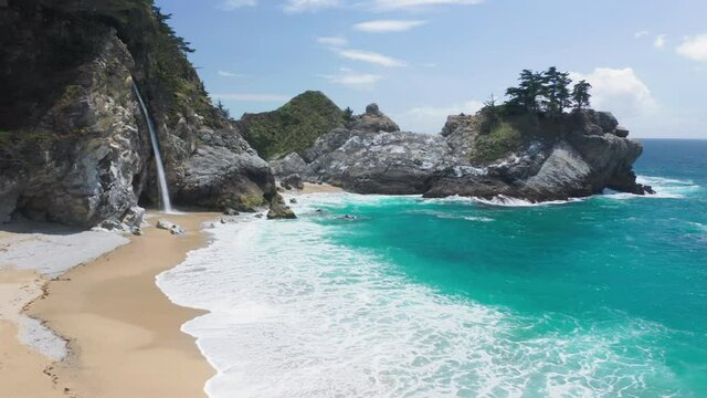 4K Aerial from the rising up perspective on the world-famous travel nature landmark on the Pacific highway &ndash; fine waterfall falling on the secluded scenic beach separated by the picturesque cliffs 