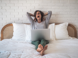 tired yawning exhausted lady in bed with laptop in Pajamas stretching her arms up at her bedroom in home office
