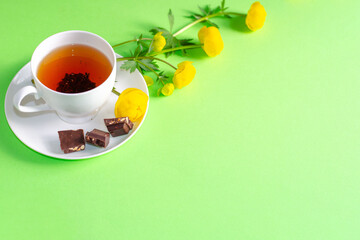 A Cup of black tea, pieces of chocolate and yellow flowers of Trollius europaeus on a green background. Sunny, bright, shadows fall from window. Chocolate day and morning tea ceremony. Copy space.