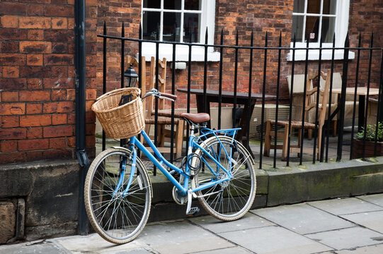 A blue bicycle with basket in the front parking in the streets leaning on a fence of a brick house