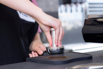 close-up photo of female hands holding a metal tamper and a portafilter with coffee in a coffee shop. A barista preparing for pressing ground coffee for brewing espresso or americano in a cafe