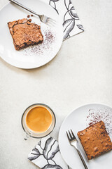 Top view of a cup of coffee and two dessert plates with brownie cake over white rustic background.