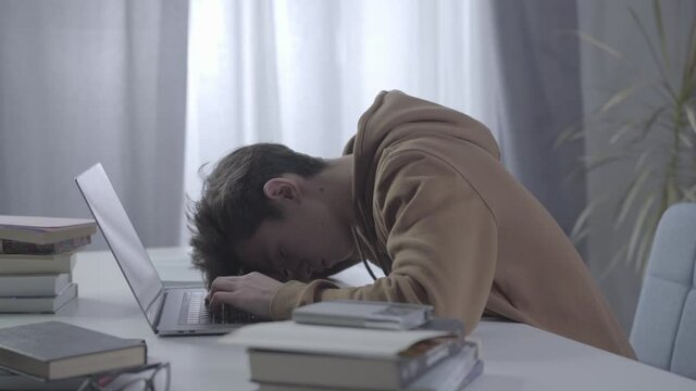 Tired college student getting asleep. Side view portrait of sleepy millennial Caucasian boy typing on laptop and putting head down on keyboard. Study hard, education, overworking.