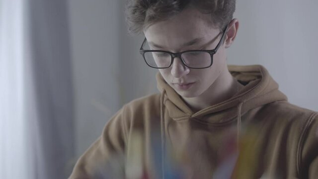 Close-up portrait of clever boy in eyeglasses doing homework. Millennial Caucasian college student holding pen and talking. Lifestyle, education, intelligence, study hard.