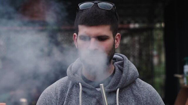 Stylish, fashionable, young guy Caucasian appearance, in sunglasses, smokes hookah from pipe, exhales, releases cloud smoke, looks camera. Happy in country, in campaign friends, against background