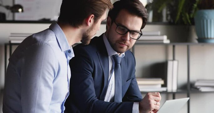 Confident businessman manager, adviser wearing suit consulting corporate client showing online project presentation on computer. Two professional business partners talking, working together in office.