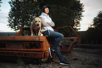 Caucasian woman in baseball cap and jeans with American Stafford dog outside near old and burnt car over beautiful sunset 