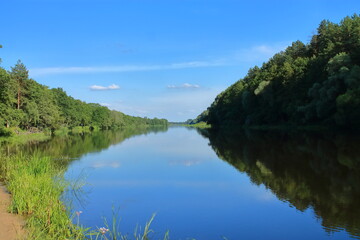 Beautiful view of the wide river from the sandy shore. Morning on the riverbank. The blue sky is reflected in the water. A great place for fishing and recreation. Banks covered with forest.