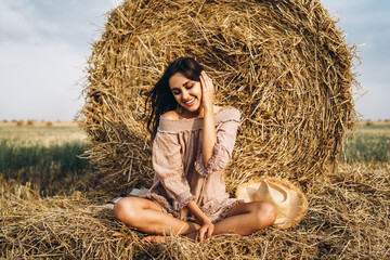Naklejka na ściany i meble A young woman with long hair and in a dress sits near a hay bale. Woman posing smiling and looking at camera