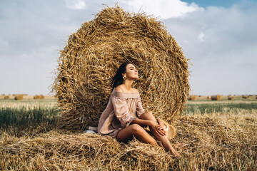 Naklejka na ściany i meble A young woman with long hair and in a dress sits near a hay bale. Woman posing smiling and looking at camera