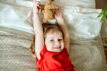 Smiling baby lies on a bed with a teddy bear in a red dress..Cute red-haired girl plays at home in a bright bedroom. funny and playful. happy childhood.Teddy bear.
