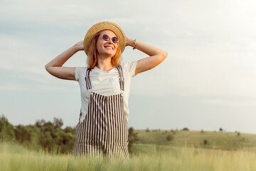 A girl in a straw hat walks on a green field at sunset. Blonde in nature on a summer day. Summer holidays.