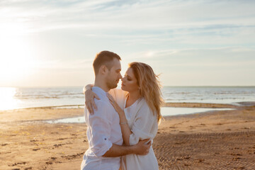 Love, sunset, romance. A young couple in long white soft clothes hugging against the backdrop of a calm sea, sunset.