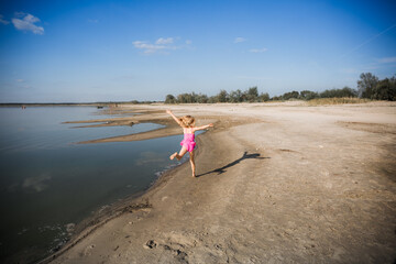 A little seven-year-old girl in a pink swimsuit runs away along the shore of the lake in a good...