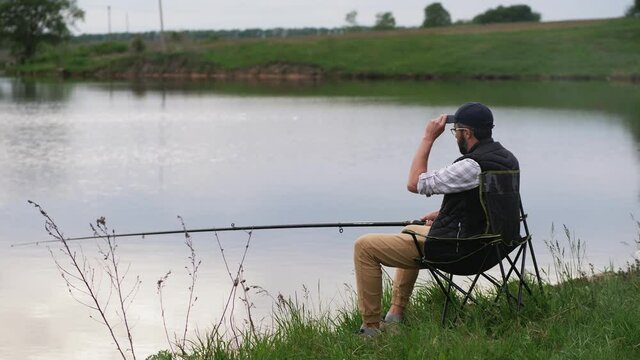 Fisherman sitting on a chair with a fishing rod in hand on the lake. A young bearded fisherman fishing on a river.