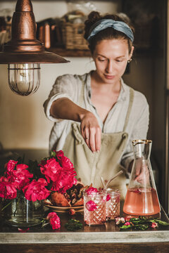 Young woman in linen apron adding ingredients to rose lemonade with ice in glasses over concrete kitchen counter with fresh pink rose flowers on it. Summer refreshing cold beverage drink concept