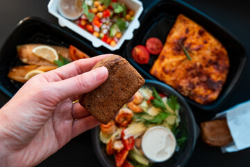 Hand holding bread on a background of food in plastic lunch boxes. Delivery service food in plastic lunch boxes on a black background. Low key, healthy food, colorful flat lay. Food concept.