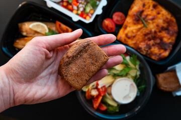 Bread in hand on a background of food in plastic lunch boxes. Delivery service food in plastic lunch boxes on a black background. Low key, healthy food, colorful flat lay. Food concept.