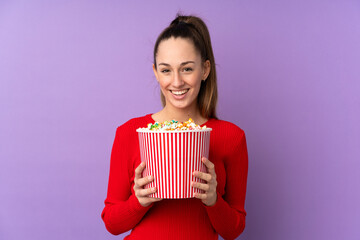 Young brunette woman over isolated purple background holding a big bucket of popcorns