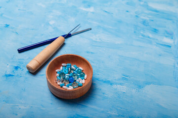 Close up of wooden bowl of beads and buttons, awl and crochet for sewing and embroidery. Set of materials for handcraft, making of bijouterie and accessories. Blue background, copy space.