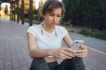 A girl in a white top and gray leggings sits in the park on a paving tile outdoors with headphones in the hands of a smartphone listening to music staring at the phone