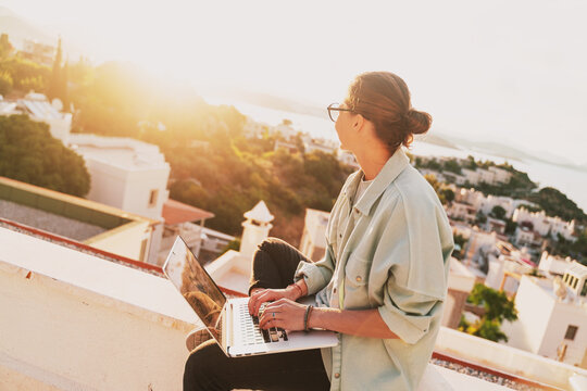 Beautiful Young Girl Woman In Eyeglasses Sitting With A Laptop On Her Balcony At Sunset With A View Of The City, Remote Work From Home, A Successful Freelancer