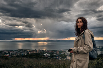 side view of a close up of a young girl with a mobile phone in hands on a hilltop with grass and cloudy sunset sky background and city view. Walking and outdoor concept.