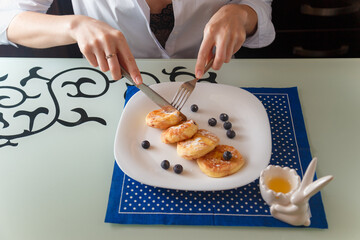 Female hands cutting cottage cheese pankaces or traditional syrniki with blueberries and honey. Healthy breakfast concept.