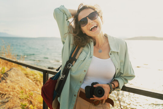 Beautiful happy with a smile young hipster girl at the sea in sunglasses with a photo camera in hand, summer vacation, travel, image retro film toning
