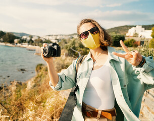 Cool beautiful happy girl woman traveler by the sea with a mask on her face, stylish outfit, with a camera in her hands, emotional expressive gestures