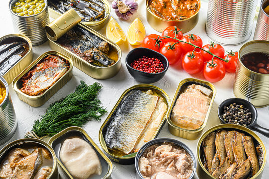 Different canned conserve products in tin cans. with fresh organic ingridients sardines, pilchard, squid, tuna pinapple, corn, peas, mango ,  beans,   over white textured background  side view