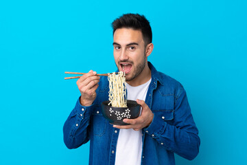 Young handsome man over isolated blue background holding a bowl of noodles with chopsticks and...
