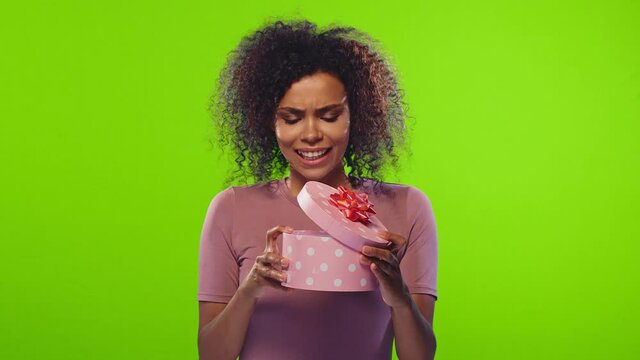 Smiling african american beautiful young woman opens gift box and is disappointed but resigned to gift in studio on green screen background. Concept of little surprises and unwarranted expectations