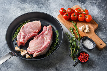Pork chop on a bone in a black frying grill pan  on a grey textured  stone background with black cloth  rosemary garlic peppercorns  and tomatoes on chopping board ingredients side  view