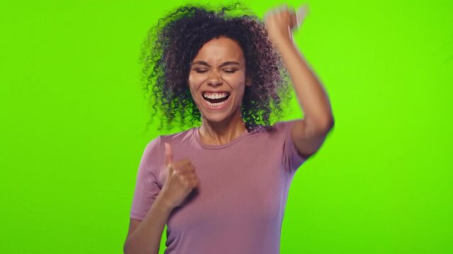 Charming cheerful beautiful african american positive woman showing thumbs up, gazes happy at camera and enjoying posing on green screen background. Positive feedback, body language concept