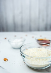 Close-up of a bowl with desiccated/dried coconut in focus with ingredients in a glass bowl on the table: maple syrup, coconut butter, and almond nuts. Concept of the homemade chocolate preparation.