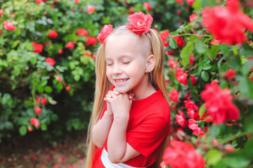 Outdoor portrait of a pretty girl of 6-7 years old against the background of a flowering red rose. A girl with long blond hair in a beautiful red dress. Little girl makes a wish closing her eyes