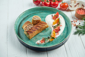 Classic Ukrainian pancakes (crepes) stuffed with meat on a blue plate with sour cream on a white wooden background. Close up view, selective focus