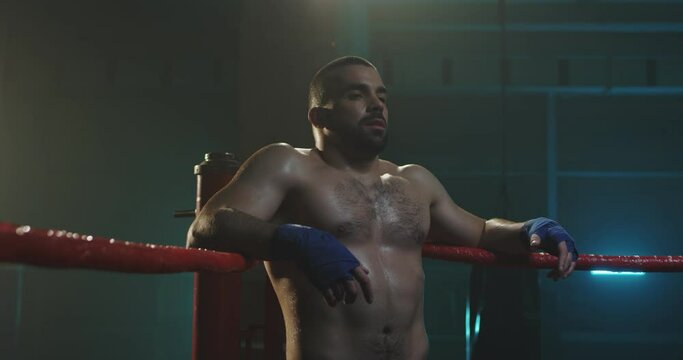Portrait of Caucasian handsome naked kickboxer sitting in corner of boxing ring in darkness and breathing hard after training. Strong male boxer having break and looking at camera. Sportsman resting.
