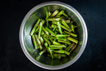 Fresh chopped green asparagus lies in a shiny bowl on a black background in the center of the composition