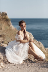 Fototapeta na wymiar Beautiful bride stands on a cliff above the sea in a glamorous white wedding dress view of veil.Romantic beautiful bride posing on the background sea. Happy bride in luxury dress posing on the beach.
