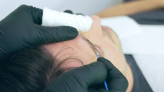 Close up of cosmetologist making permanent makeup. Microblading procedure. High quality close-up 4k slow motion video of a man laying on a couch during the esthetician brow microblading treatment.