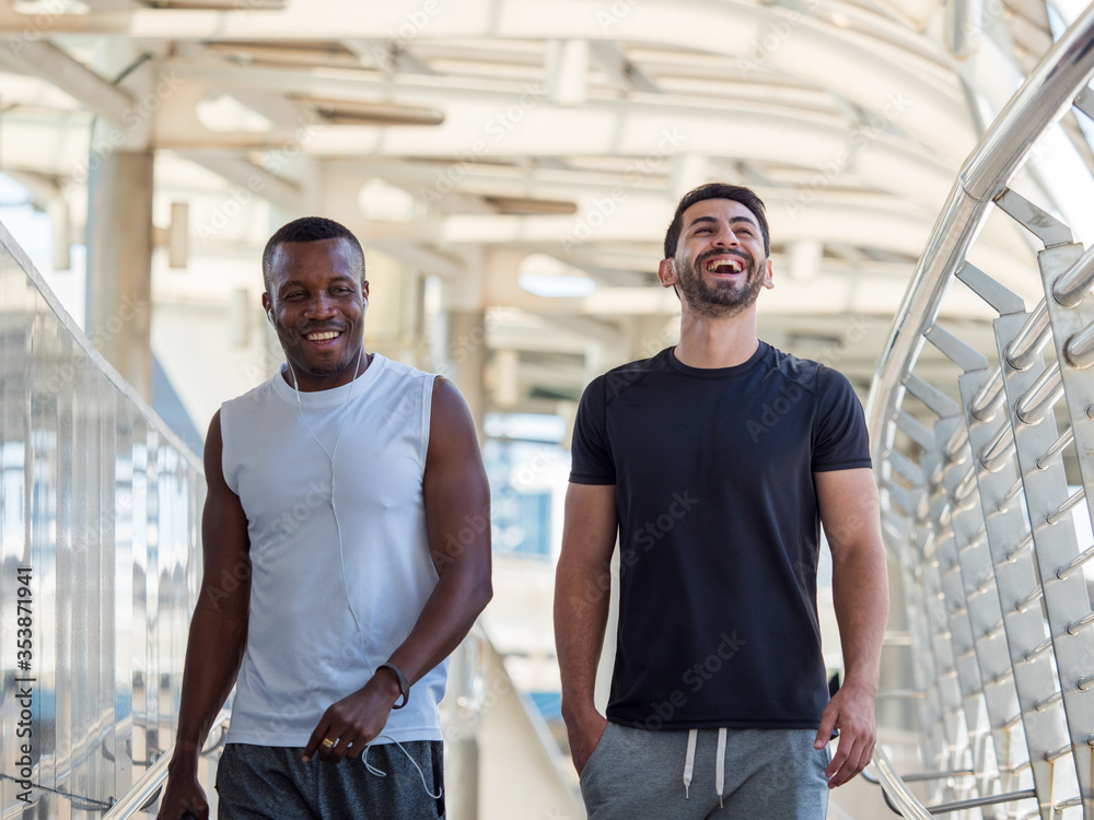 Wall mural two diversity male athletes laughing while walking under the shade after jogging in the city.