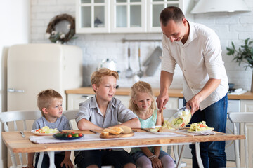 family father with three kids two sons and daughter eating healthy food in kitchen at home, dad puts green salad on plates to his kids.
