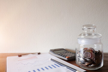 The cropped view of the glass jar contains the coins withe calculator and business paper on the wood desk with white natural blackground for texing the message, information,and background.