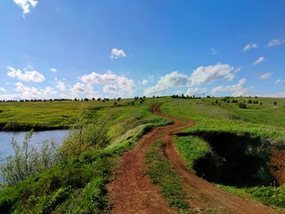 Obraz premium rural winding road uphill through a field between a lake and a ravine against a blue sky with clouds on a sunny day